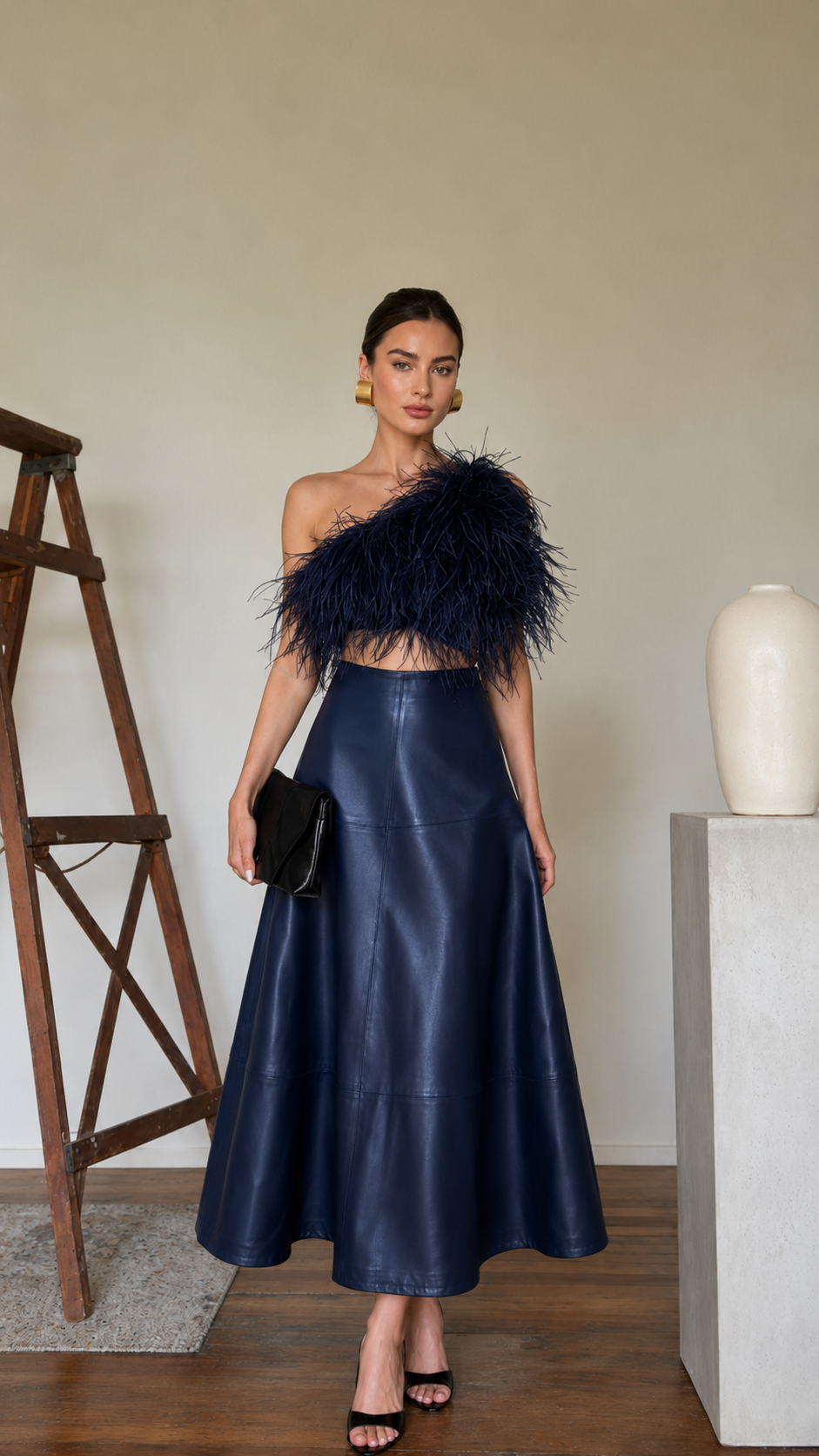 Model wearing a navy high-waisted faux leather A-line midi skirt with a statement one-shoulder feather top