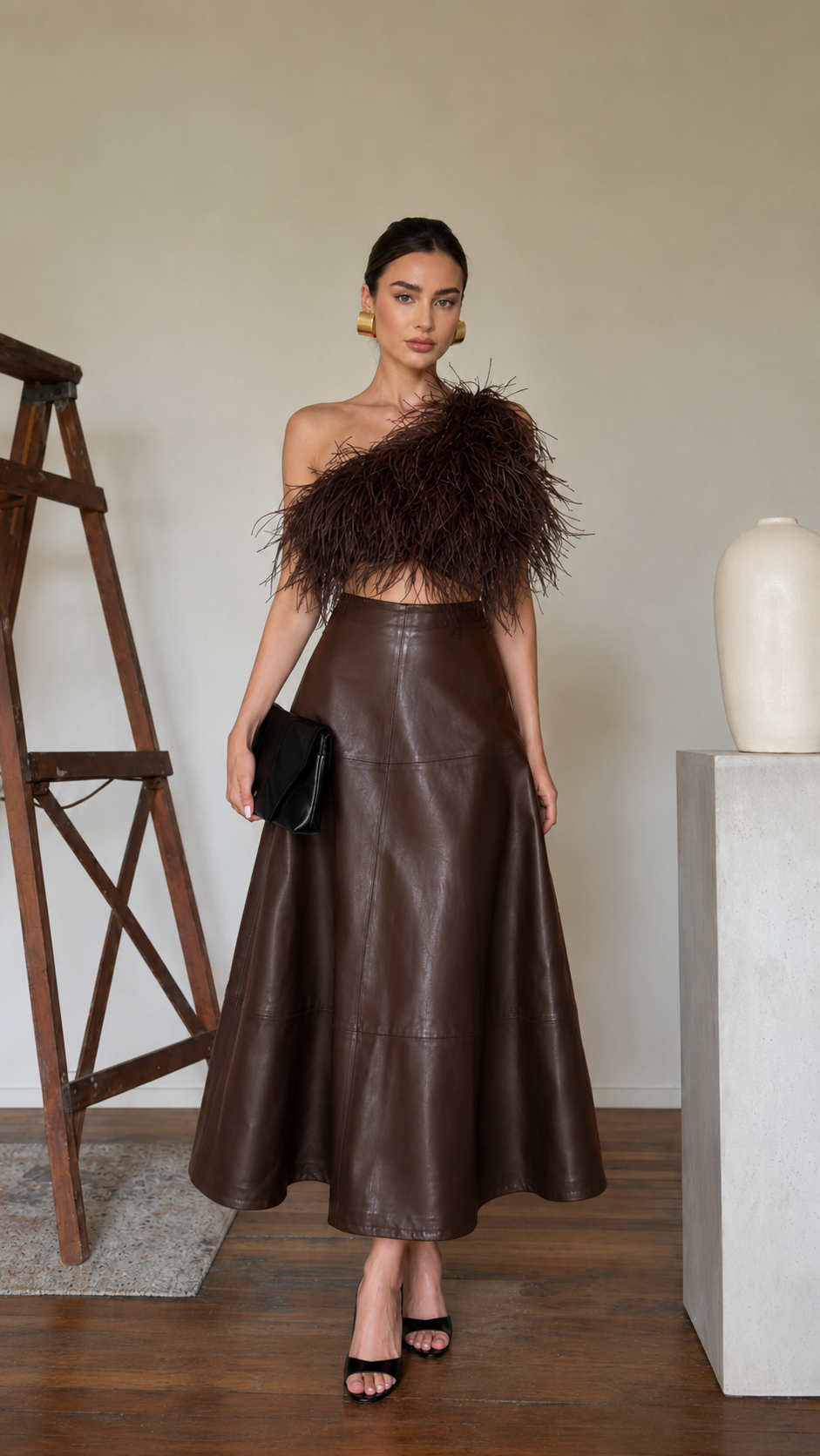 Model wearing a chocolate brown high-waisted faux leather A-line midi skirt with a one-shoulder feather top