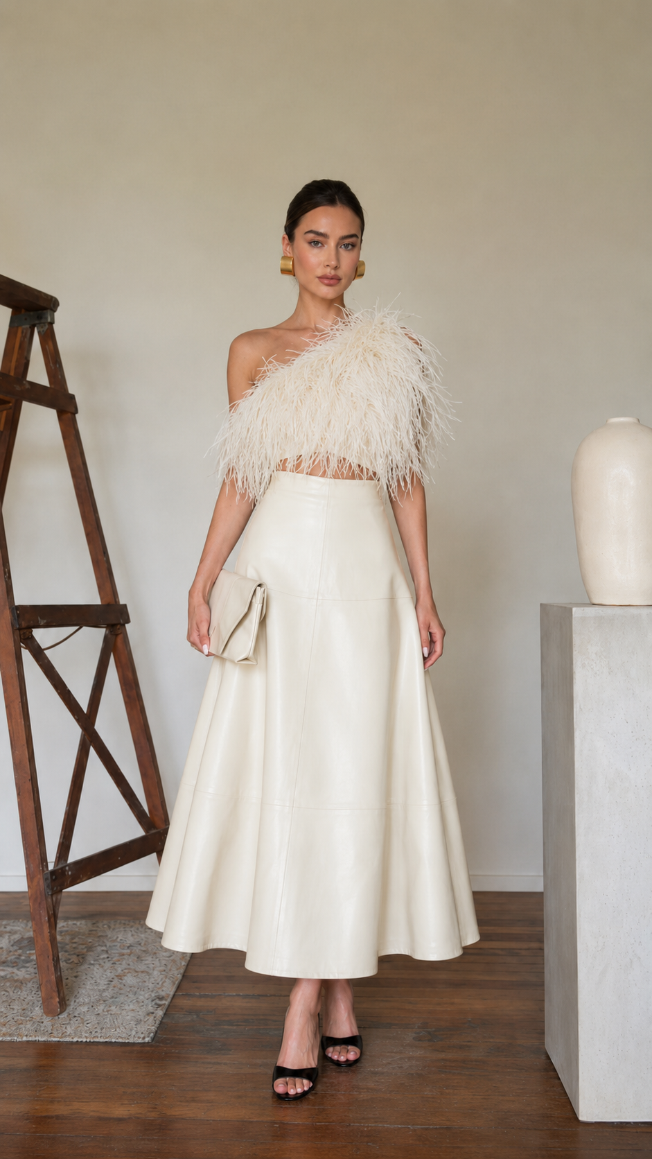 Model wearing an ivory high-waisted faux leather A-line midi skirt with a feather top and clutch